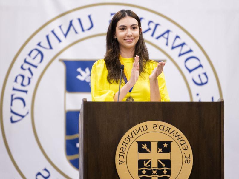 The image shows a woman standing at a podium with the logo of "St. 爱德华。's University" in the background. She is wearing a bright yellow top and is clapping with a smile on her face. The podium has the university's emblem on the front. She appears to be addressing or acknowledging an audience, possibly during a formal event or ceremony at the university.