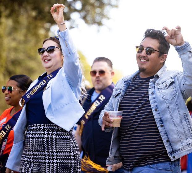 Two alumni walk in the homecoming parade with fellow alumni and make a toppers up hand sign.