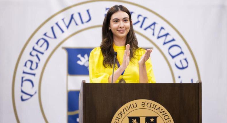 The image shows a woman standing at a podium with the logo of "St. Edward's University" in the background. 她穿着一件亮黄色的上衣，面带微笑地鼓掌. The podium has the university's emblem on the front. She appears to be addressing or acknowledging an audience, possibly during a formal event or ceremony at the university.