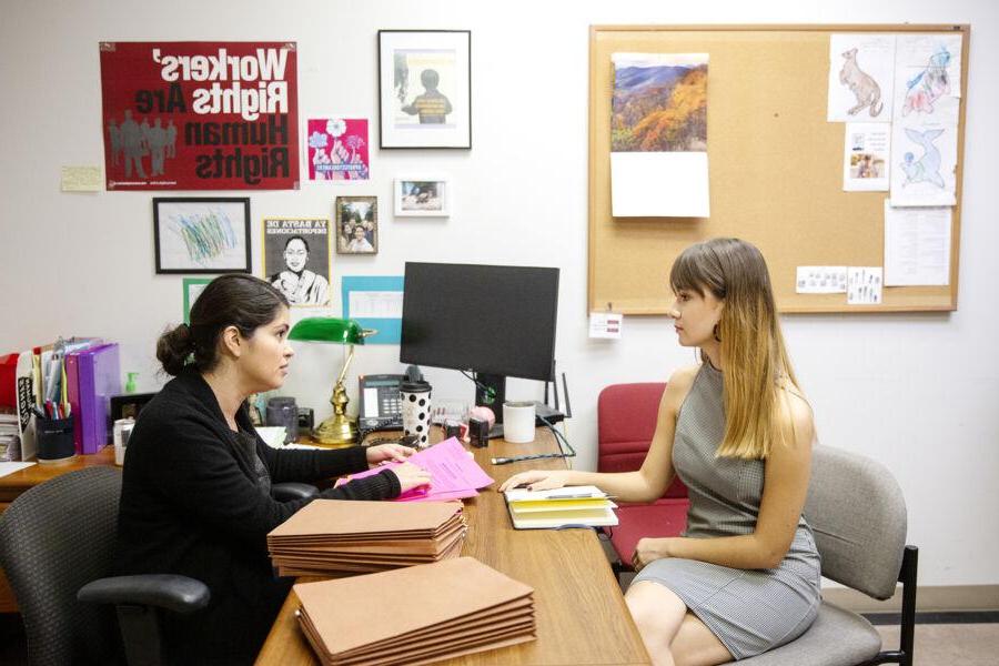 The image depicts two women in an office setting. 一个留着浅棕色长发的女人穿着一件无袖的灰色连衣裙，坐在办公桌前. She is engaged in conversation with another woman, who has dark hair tied back and is wearing a black top. 第二个女人拿着粉红色的文件，桌上堆着几个棕色的文件夹. 办公室的墙上装饰着各种各样的海报和图画. A corkboard with drawings and notes is also visible.