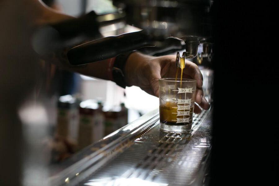 The image shows a close-up of a barista's hand pulling a shot of espresso using an espresso machine. The coffee is being dispensed into a small, clear measuring glass with markings indicating fluid ounces. 场景设置在咖啡厅的环境中, 重点是富人, 浓咖啡斟满了杯子, capturing the process of coffee preparation.